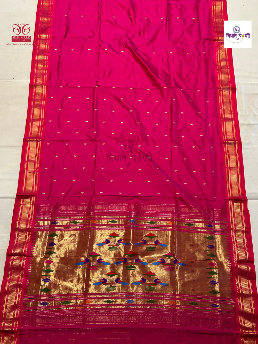 A bright pink silk saree with a wide gold zari border, small metallic dot motifs, and a decorative gold brocade pallu with colorful thread embroidery, laid flat on a light background.