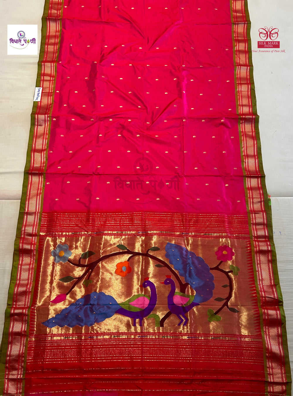 A bright pink and red silk saree with a wide gold zari border, featuring embroidered peacock motifs in blue, pink, and green on the pallu section.