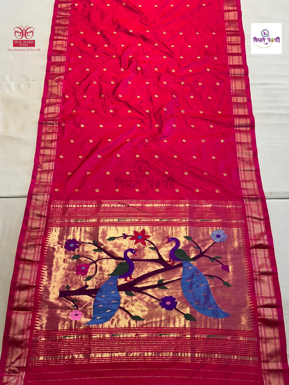 A bright pink silk saree with a wide gold zari border, decorated with two blue peacocks and floral motifs on the pallu.