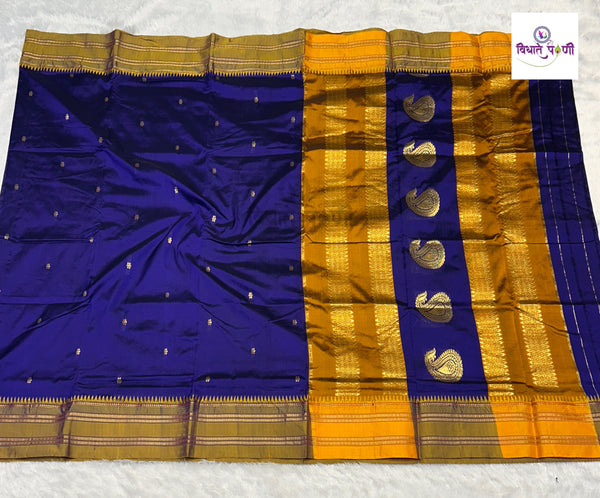 A royal blue and gold zari silk saree laid flat on a white textured surface, featuring a wide gold border, a vertical gold stripe with metallic paisley motifs, and small gold dots on the blue body.