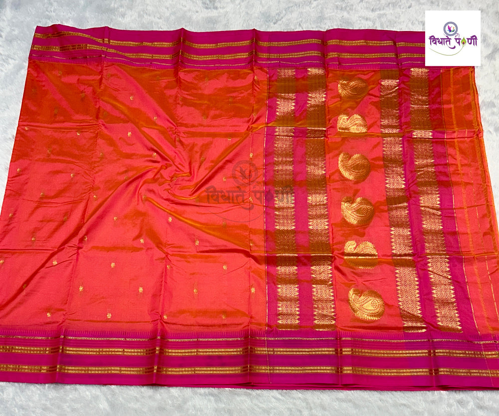 A bright pink and orange-red silk saree with intricate gold zari weaving, laid flat on a light gray textured surface. The fabric features a wide border with gold stripes and a vertical section decorated with gold paisley motifs.