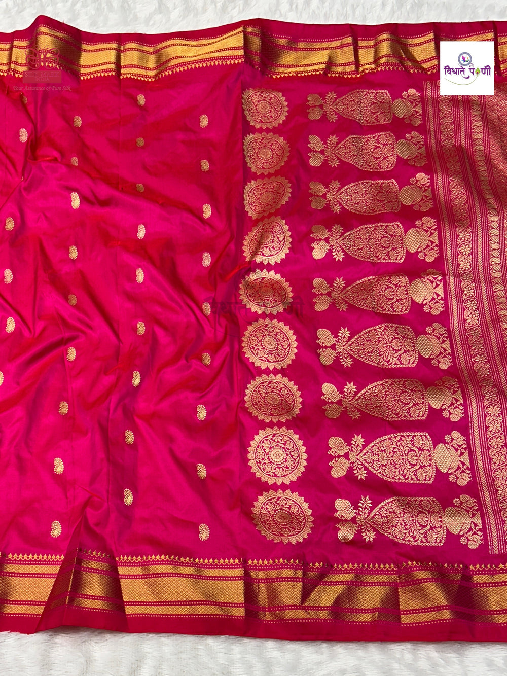 A bright pink silk saree with intricate gold Banarasi zari embroidery, featuring a wide border with floral motifs and a small brand label in the top right corner.