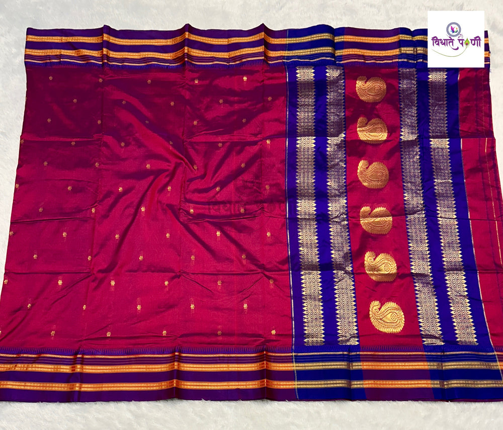 A folded and spread-out Indian silk saree in magenta pink with a royal blue border, gold zari metallic patterns, and small gold motifs, displayed on a white textured background.