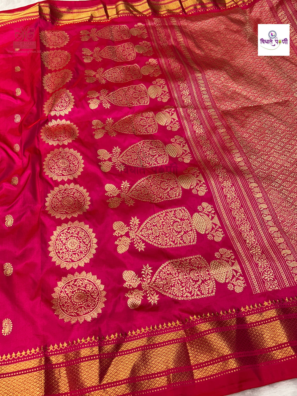 A close-up of a bright pink silk saree with intricate gold Zari woven patterns, featuring paisley motifs, floral mandalas, and a decorative border.