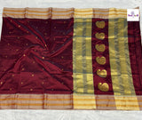 A deep maroon silk saree with gold zari borders, a vertical band of gold paisley motifs, and small gold dots on the body, laid flat on a white textured surface.