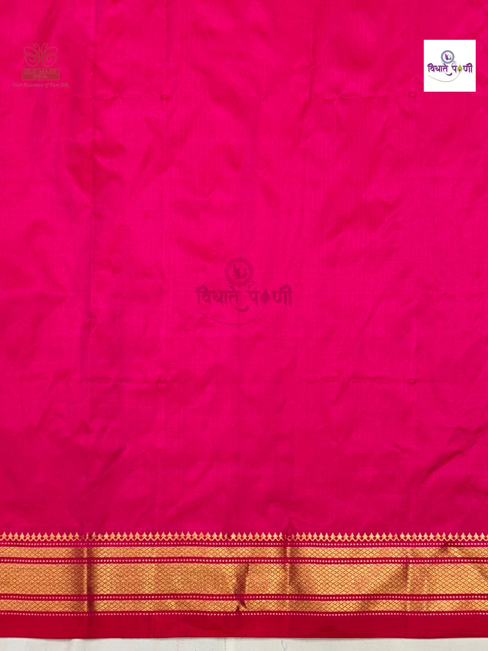 A bright pink silk saree fabric with a gold and red border at the bottom, featuring a small white brand label in the top right corner.