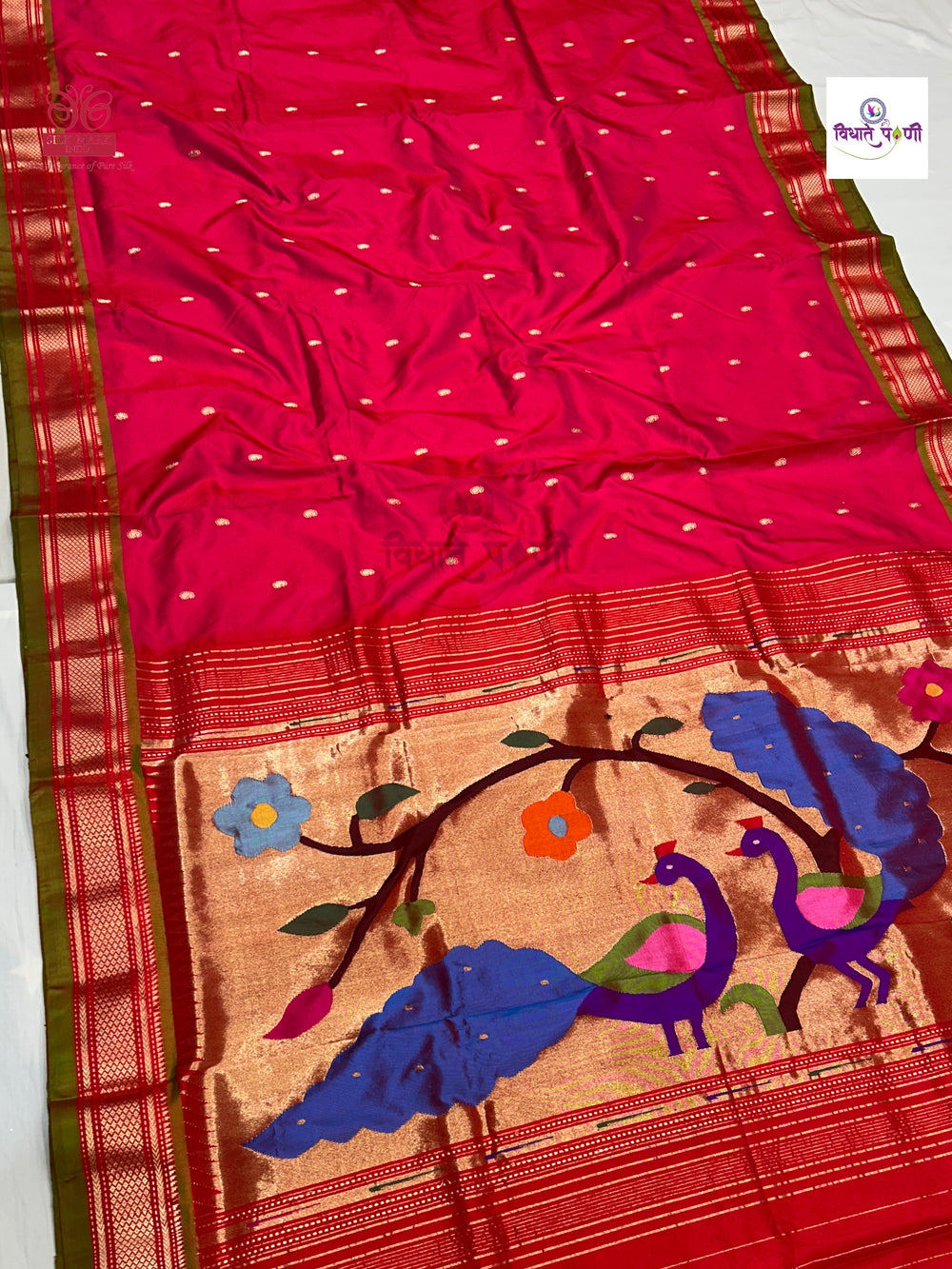 A red and gold Banarasi silk saree with a wide gold zari border, small gold dot motifs, and a detailed handwoven floral and peacock pattern on the pallu.